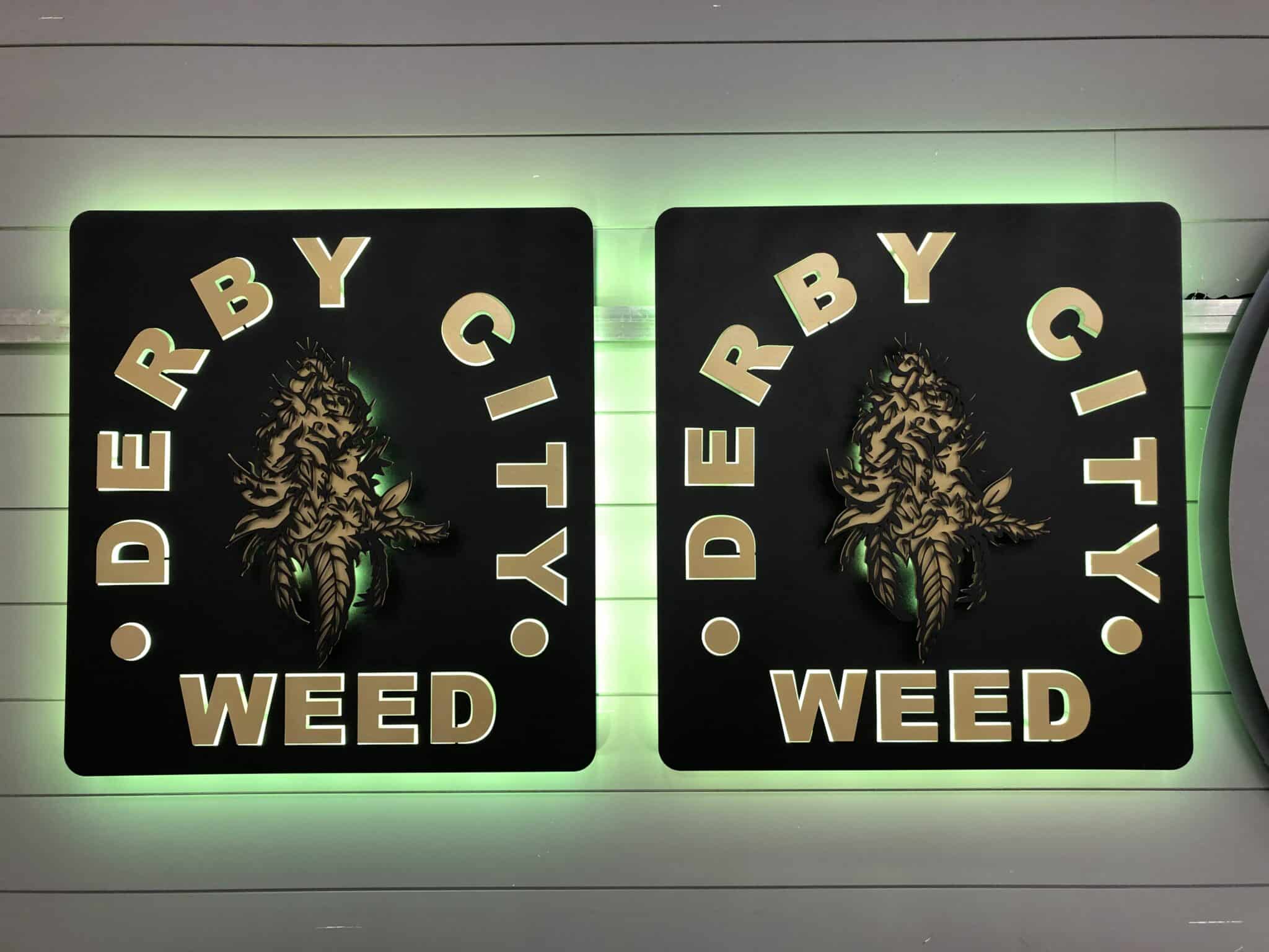 Two Derby City Weed square signs side by side, gold cannabis leaves on black with a green LED halo.