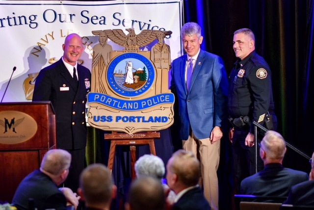 Portland Police and Navy officers at a podium beside a presented USS Portland seal.