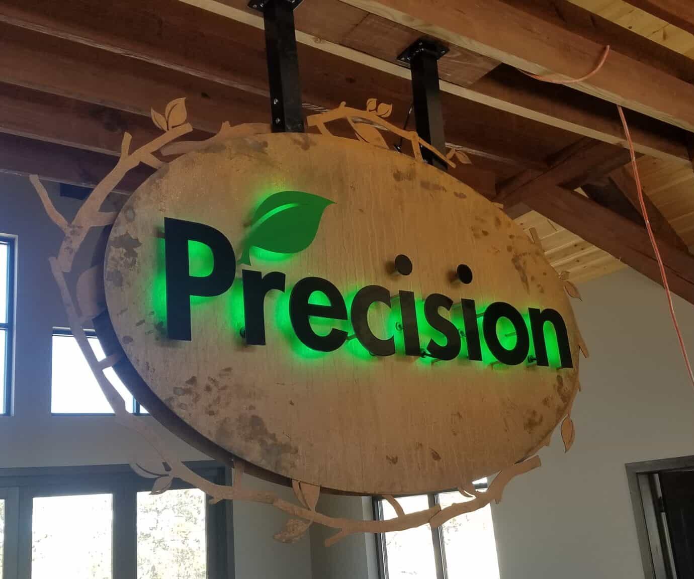 Precision Landscaping oval sign hanging from a beam, green lit lettering with a leaf on a wood face.