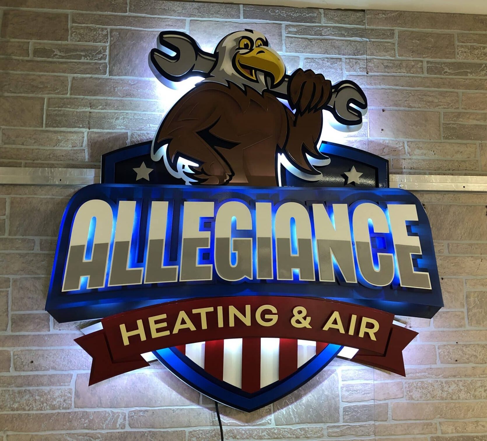Allegiance Heating & Air shield sign with an eagle holding a wrench, halo-lit LED on brick.