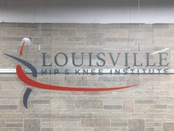 Louisville Hip & Knee Institute acrylic sign with dimensional letters above a red and gray swoosh, mounted on stone.