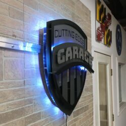 Cutting Edge Garage shield edge-lit sign in black with louvered slots,  glowing blue.