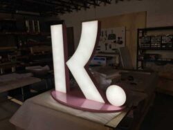 Freestanding fabricated face-lit Klarna K sign on its pink base in the shop, standing taller than the bench beside it.