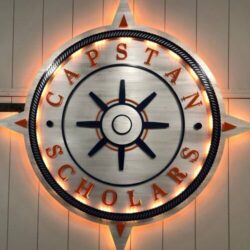 UW Platteville Capstan Scholars sign, a brushed ship's wheel inside a rope ring, glowing warm amber.