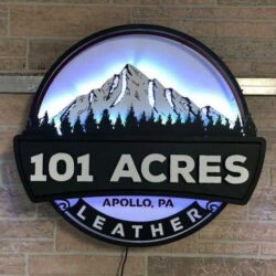 101 Acres Leather sign with a snow-capped mountain and pine tree skyline, its LED halo glowing white against exposed brick.