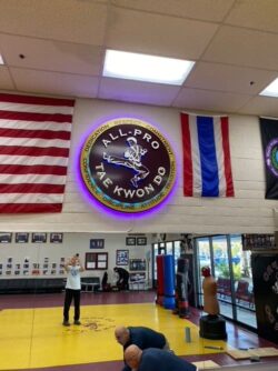 All-Pro Tae Kwon Do halo-lit sign installed high on the dojo wall above the mats and flags, glowing purple.