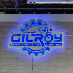 Gilroy Motorcycle Center sign with the name across a cut gear and piston, glowing blue.