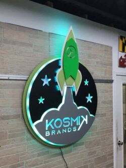 Kosmik Brands round sign with a green rocket rising past stars, halo-lit LED and glowing teal.