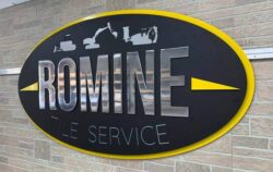 Romine Tile Service oval sign in black with a yellow rim, polished aluminum lettering under cut machinery silhouettes.
