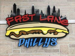 Fast Lane Phillys sign with a loaded cheesesteak beneath the name and a city skyline above it.