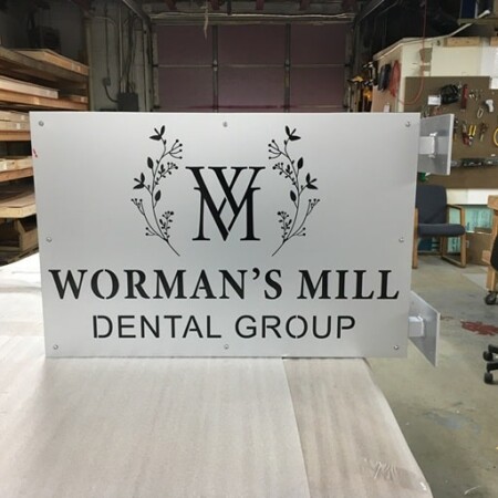 Worman's Mill Dental Group blade sign face down in the shop, a WM monogram framed by botanical sprigs.