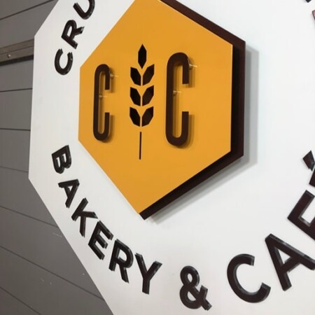 An up-close shot of Crust & Crumb Bakery & Café hexagonal blade sign in white, with a yellow hexagon monogram and a wheat stalk.
