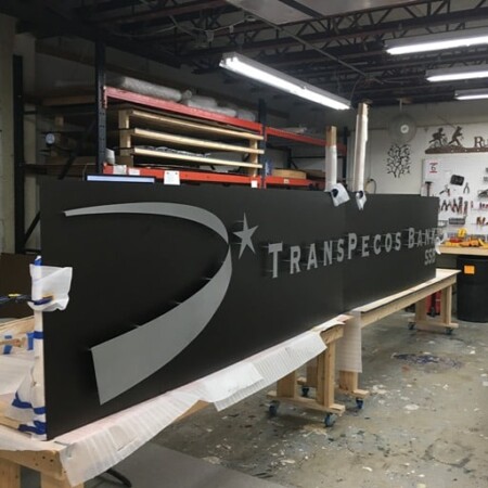 TransPecos Banks sign on the bench in the shop, brushed lettering and a swoosh across a long charcoal panel.