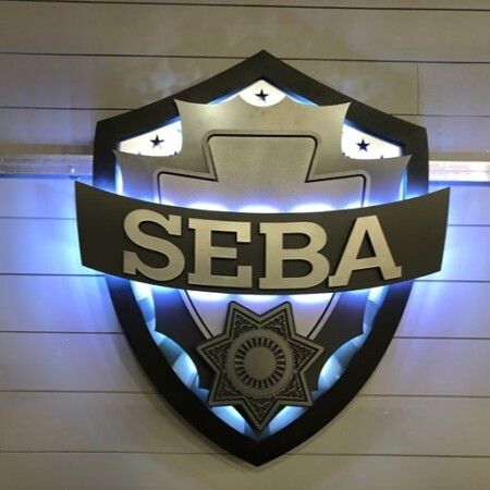 SEBA shield sign in black and gold, halo-lit LED and glowing blue behind the banner.