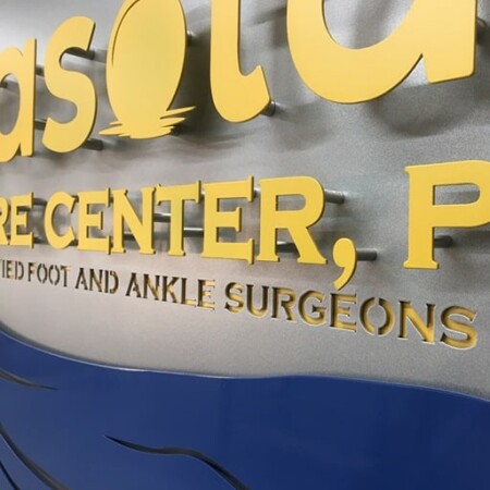 Close view of the Sarasota Foot Care Center letters, yellow faces standing off the brushed disc on studs.
