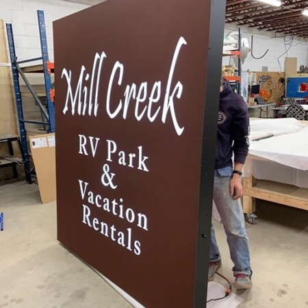 The Mill Creek RV Park face-lit lightbox seen edge-on with a fabricator beside it, taller than a person.
