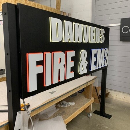 Danvers Fire & EMS panel sign on the bench in the shop, white and red letters outlined on black.