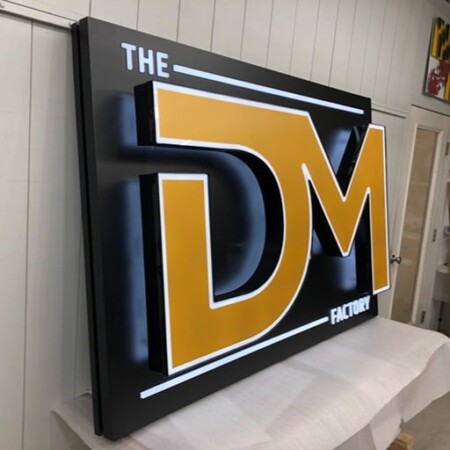 The DealMaker Factory panel sign on the bench in the shop, gold DM letters on black inside a white outline.
