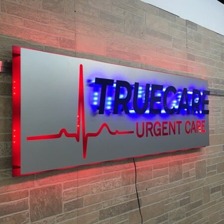 TrueCare Urgent Care sign with a red heartbeat line running under the name, glowing red and blue on brick.