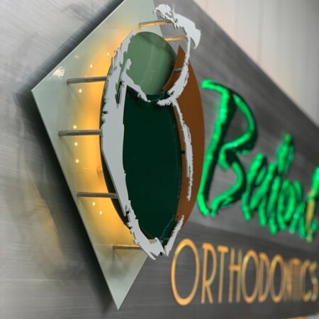 Bedont Orthodontics halo-lit LED panel sign in brushed aluminum, green script over copper lettering, glowing warm white.