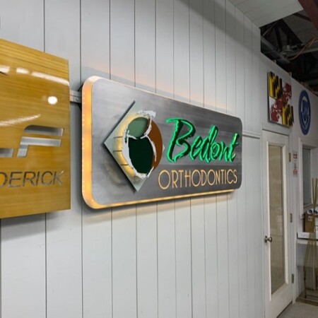 Bedont Orthodontics halo-lit LED panel sign in brushed aluminum, green script over copper lettering, glowing warm white.