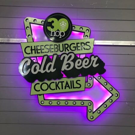 30 Hop cheeseburgers, cold beer and cocktails arrow sign in olive green and black, its LED halo glowing magenta on a gray panel wall.