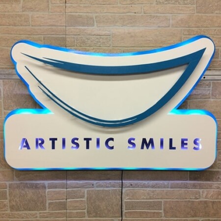 Artistic Smiles dental sign shaped as a smile in cream and blue, its LED halo washing the brick behind it.