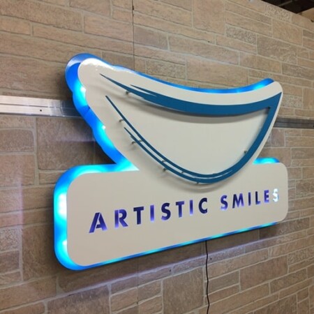 Artistic Smiles smile-shaped dental sign angled so the blue LED halo behind the panel reads clearly.