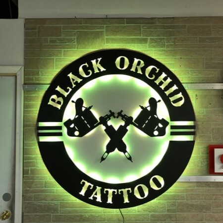 Black Orchid Tattoo Studio halo-lit LED round sign with crossed tattoo machines, glowing green on brick.