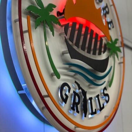 Gulf Shore Grills sign from an angle, the layered grill and palms lit blue from behind.