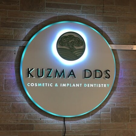 Kuzma DDS Cosmetic and Implant Dentistry round sign in white, halo-lit LED and glowing cyan on brick.