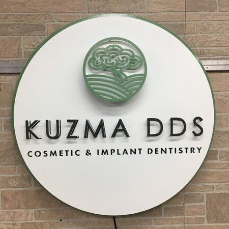Kuzma DDS Cosmetic and Implant Dentistry round sign in white with a green emblem, mounted on stone.