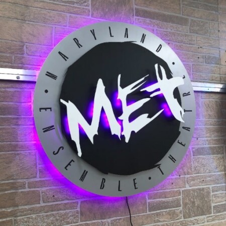 Maryland Ensemble Theater round sign with MET in white brushstrokes, halo-lit LED and glowing purple.