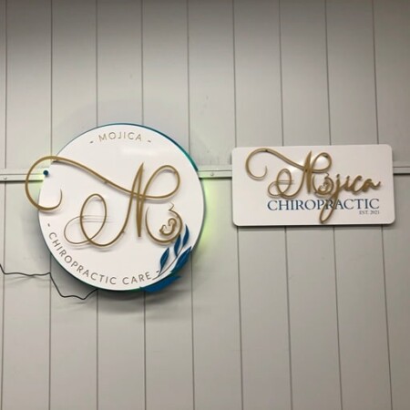 Mojica Chiropractic Care round sign with a gold monogram, beside a smaller wooden nameplate on the same wall.
