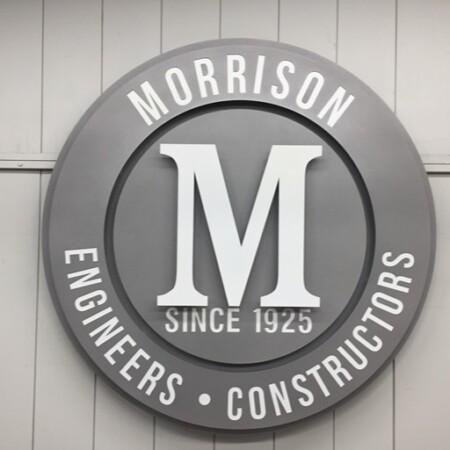 Morrison Engineers & Constructors round sign in gray with a raised M, Since 1925 beneath it, photographed unlit.