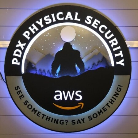 PDX Physical Security round sign with a bigfoot silhouette under the stars above the AWS mark, glowing blue.