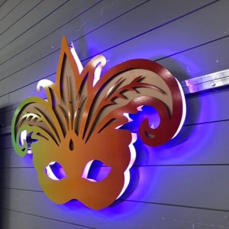 Boudreaux Cajun Steakhouse halo-lit LED Mardi Gras mask sign in a green-to-red gradient, glowing blue against a paneled wall.