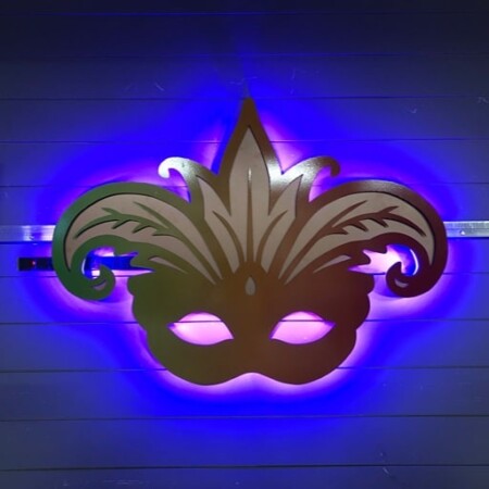 Boudreaux Cajun Steakhouse halo-lit LED Mardi Gras mask sign in a green-to-red gradient, glowing blue against a paneled wall.