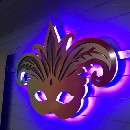 Boudreaux Cajun Steakhouse halo-lit LED Mardi Gras mask sign in a green-to-red gradient, glowing blue against a paneled wall.