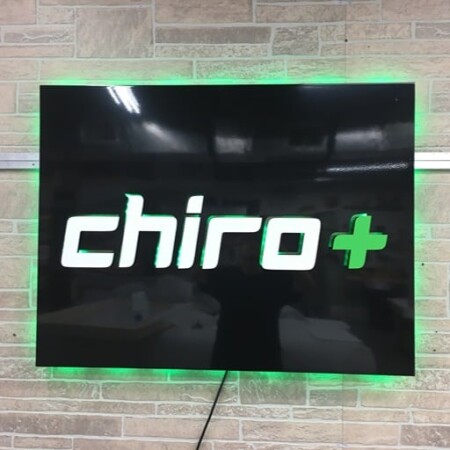 Chiro Plus halo-lit LED panel sign in gloss black with white lettering and a green cross, glowing green on stone.