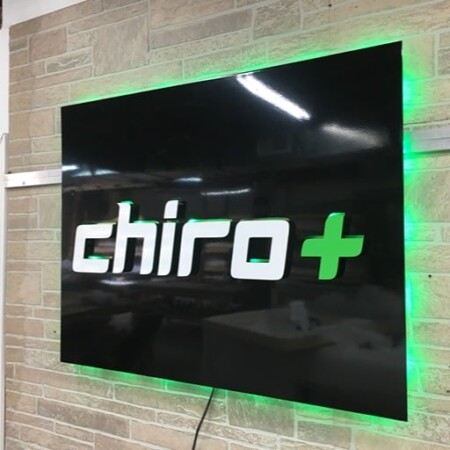 Chiro Plus halo-lit LED panel sign in gloss black with white lettering and a green cross, glowing green on stone.