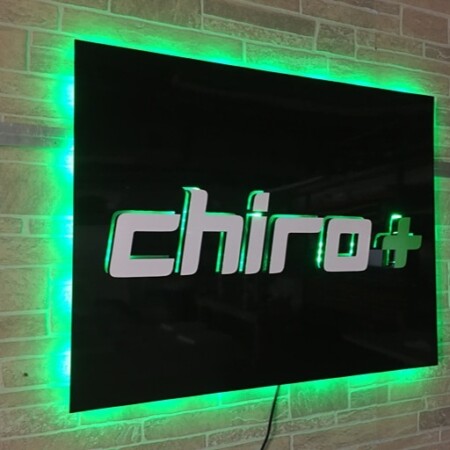 Chiro Plus halo-lit LED panel sign in gloss black with white lettering and a green cross, glowing green on stone.