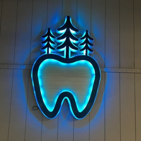 BH Dentures and Implants halo-lit LED sign shaped as a tooth beneath pine trees, glowing cyan.