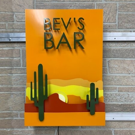 Bev's Bar panel sign with a layered desert sunset and cacti, the name cut into an orange face, mounted on brick.