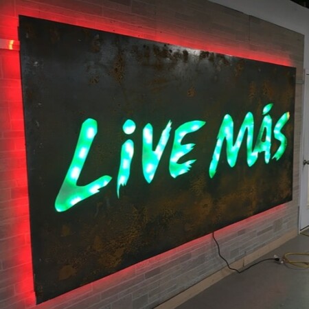 Live Más cut straight through a rusted steel panel, the letters glowing green inside a red halo.