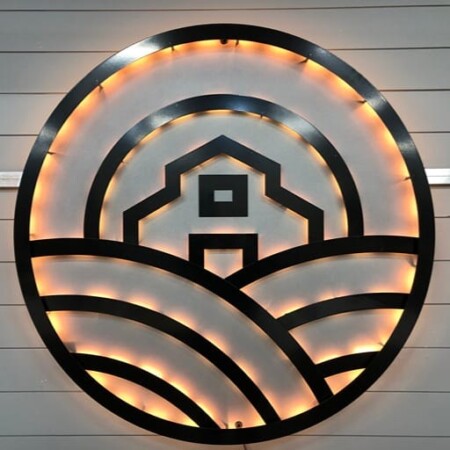 Pono Acres round sign, a barn and field cut in matte black with a warm white LED halo behind.