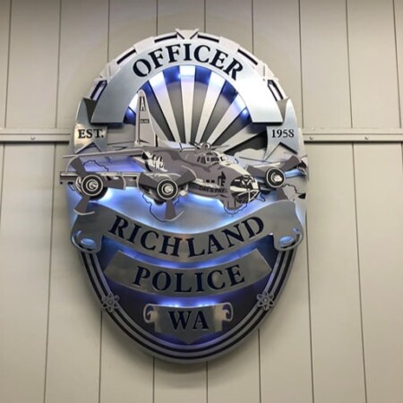 Richland Police badge sign in layered steel with patrol car and aircraft detail above a name banner, glowing blue.