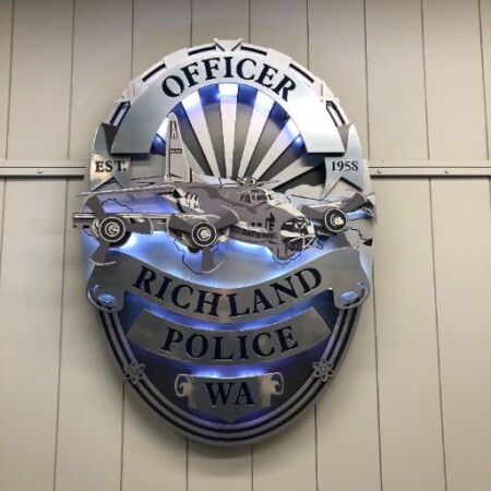 Richland Police badge sign in layered steel with patrol car and aircraft detail above a name banner, glowing blue.