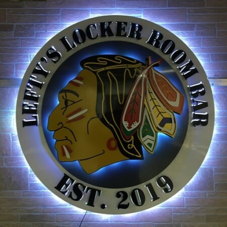 A circular halo-lit led sign with an Indian head in the center, reading Lefty's Locker Room Bar. It is back-glowing white against a stone wall.