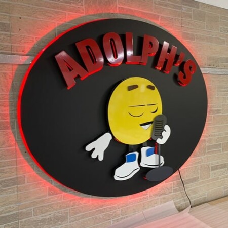 Adolph's Grill and Bar round metal sign with a cartoon lemon character, lit by red halo lighting.